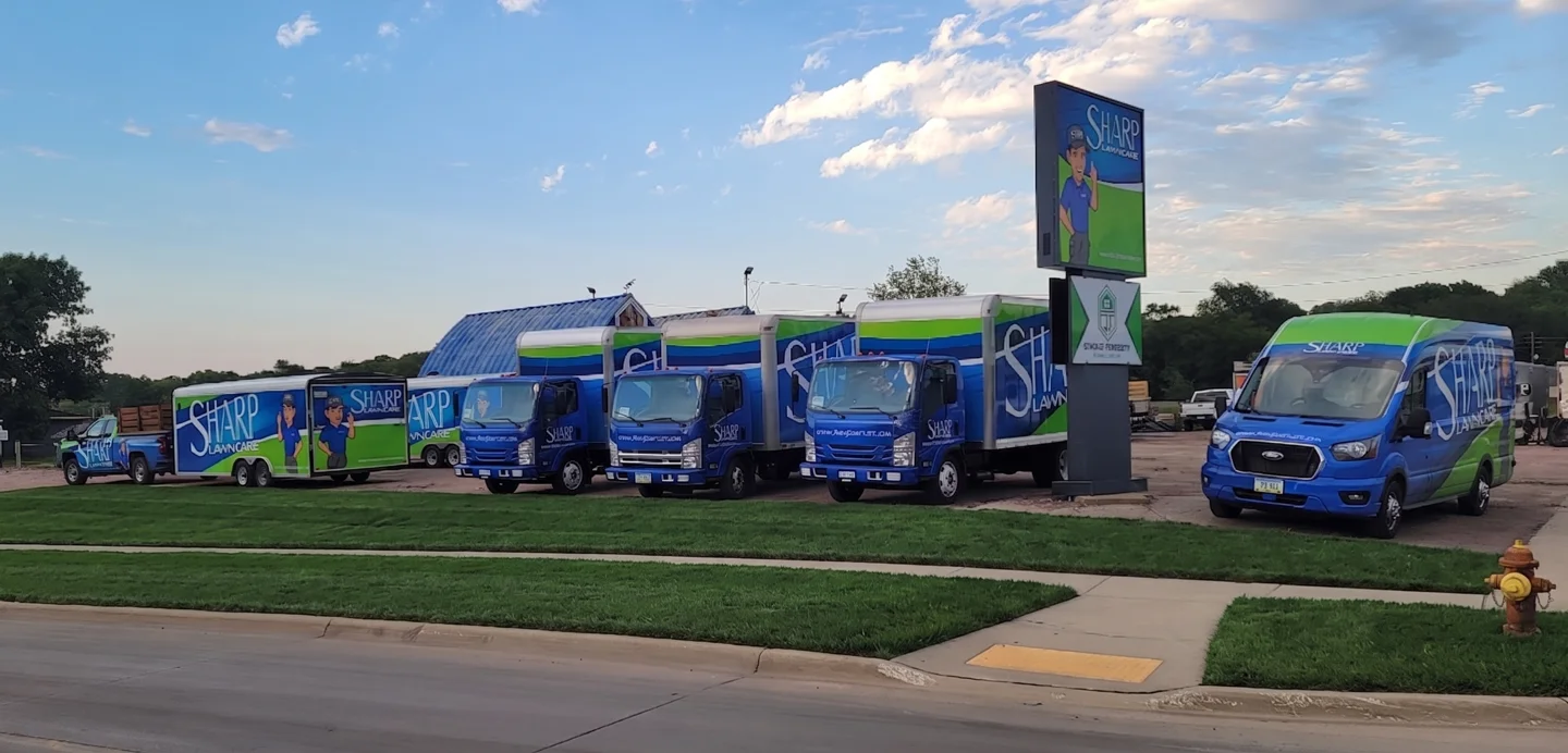Sharp Lawn Care fleet of branded box trucks, van, and pickup parked at our Sioux City branch on 20th Street with the illuminated mascot sign visible behind the trucks.