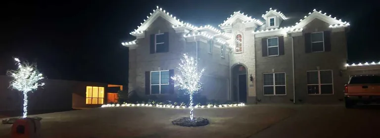 Cool white Christmas lights on every gable of a stone two-story home with two bare trees wrapped to the branch tips