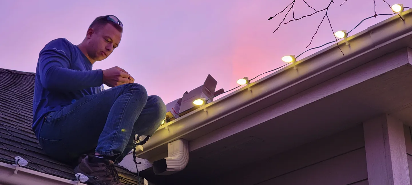 A Sharp Lawn Care technician on a roof edge clipping warm white bulbs to the gutter line at sunset during a Christmas light installation
