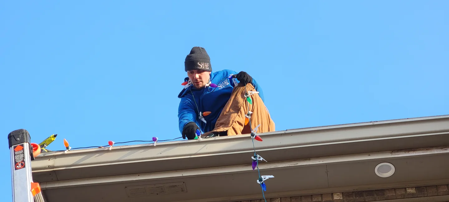 A Sharp Lawn Care installer pulling a multicolor Christmas light strand along a roofline against a clear winter sky