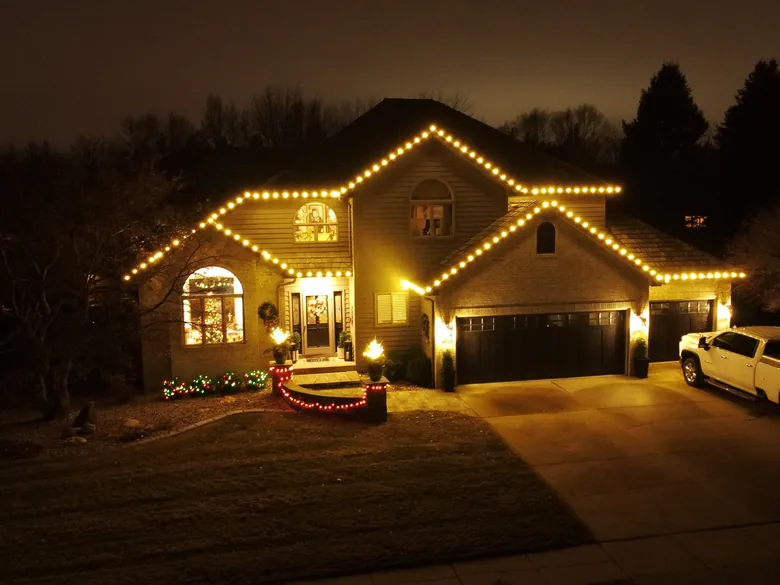 A two-story home at blue hour with warm white Christmas lights along every roofline, a lit wreath and shrubs at the entry and a pickup in the drive
