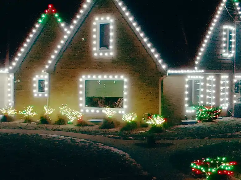 A home at night with warm white lights outlining the steep gables and every window, red bulbs at the roof peaks, and lit shrubs across the front yard