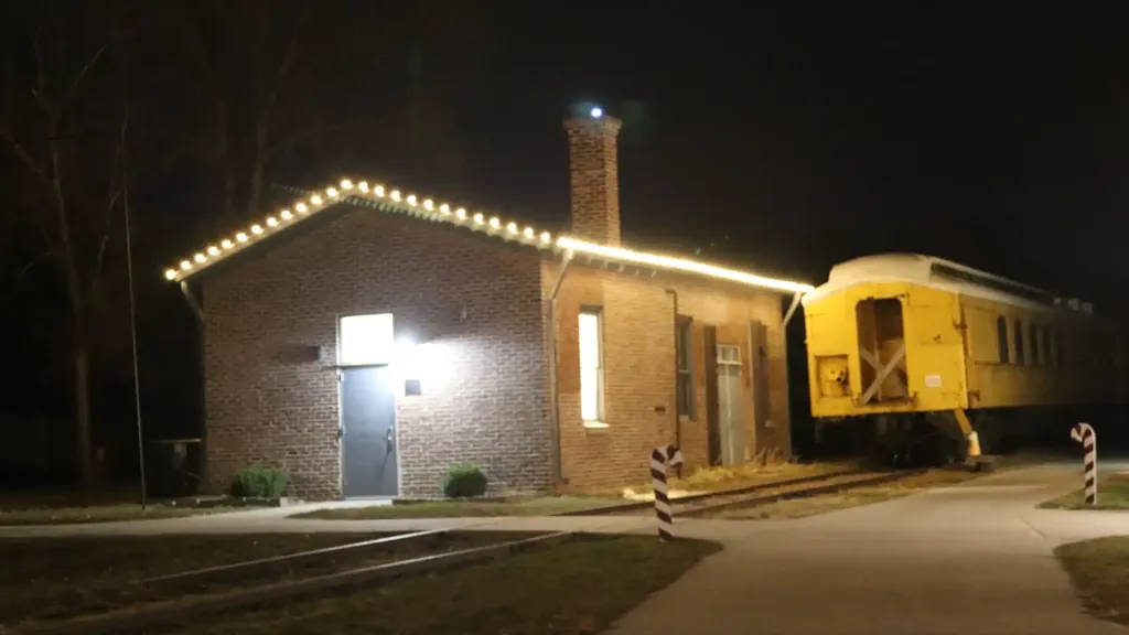 The brick depot at the Sioux City Railroad Museum lit along the roof line, with a yellow rail car on the track alongside