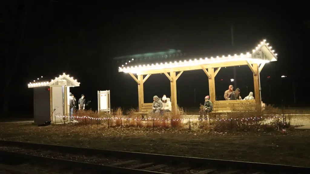 A timber shelter and a small station building at the Sioux City Railroad Museum edged in lights, with low strings along the beds