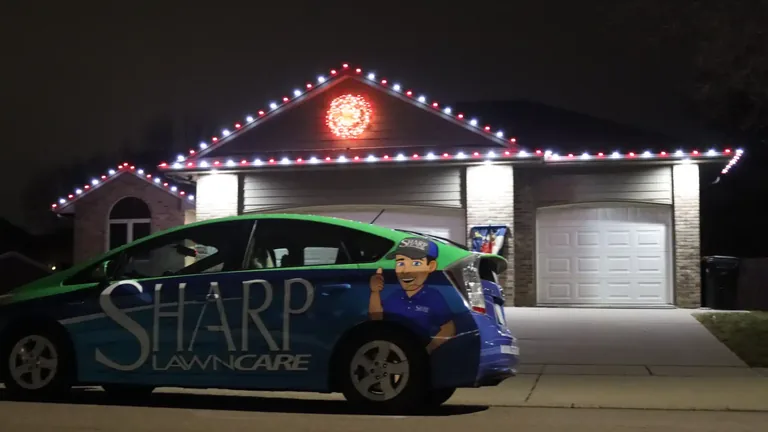 Red and white Christmas lights along the full roofline of a brick ranch home at night, a lit wreath in the gable and the Sharp car parked in the driveway