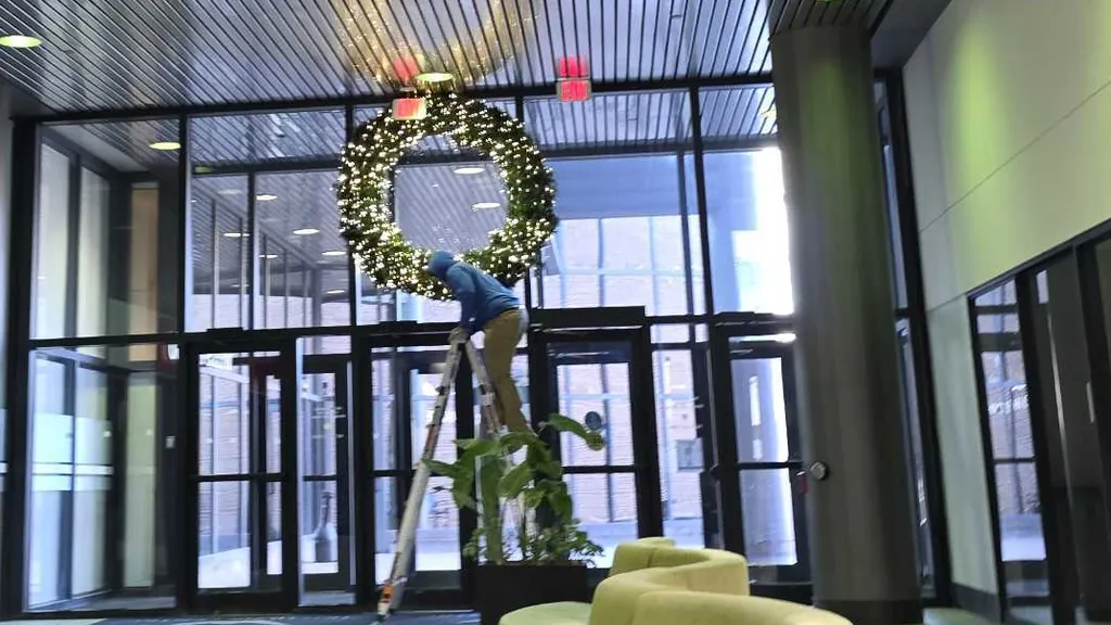 A Sharp Lawn Care installer on a stepladder hanging a large lit wreath above the glass entrance of a commercial lobby