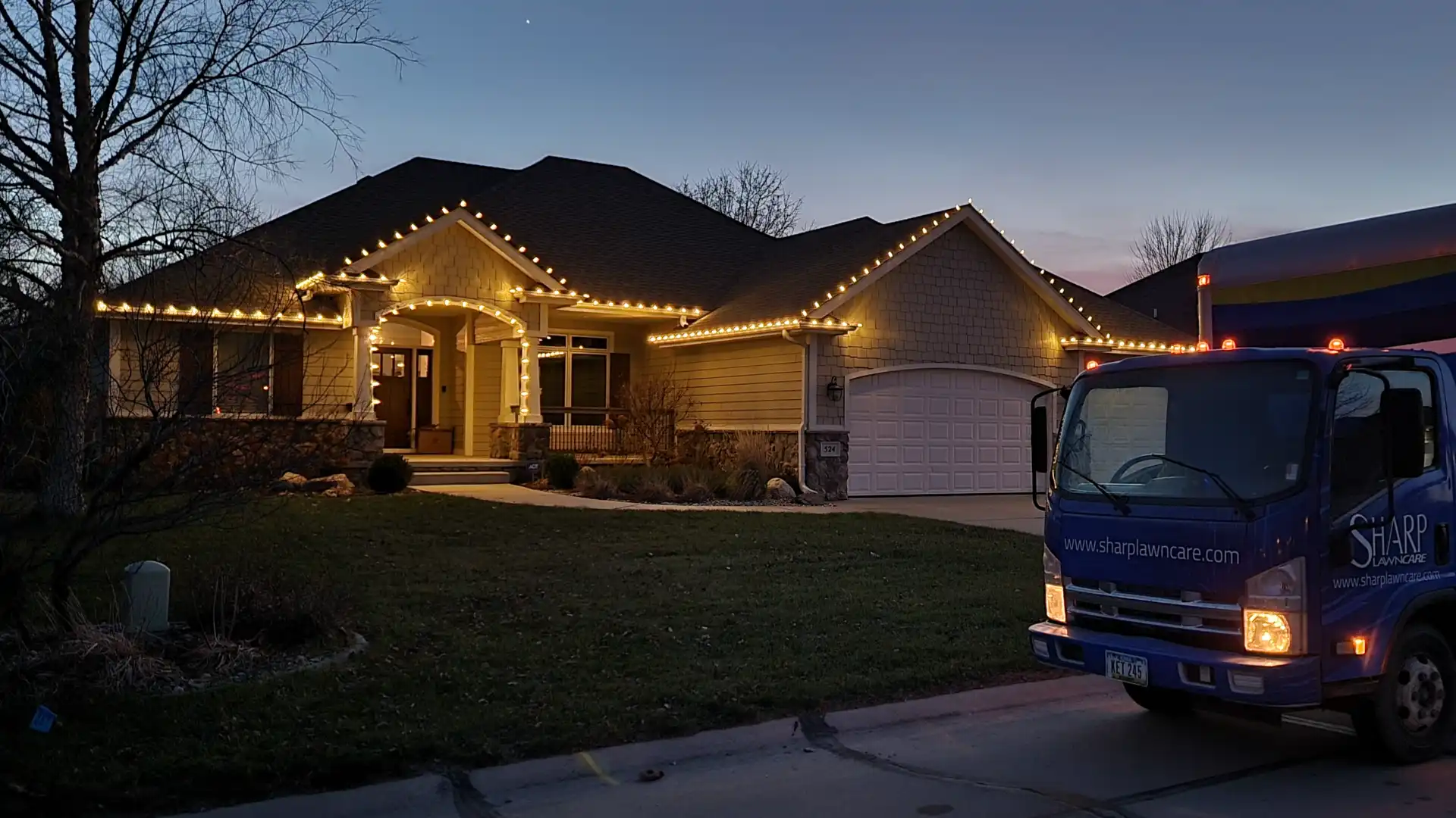 A house in Sioux City, Iowa, professionally decorated with warm white Christmas lights along its roofline and porch, with a Sharp Lawn Care truck in the driveway at dusk.