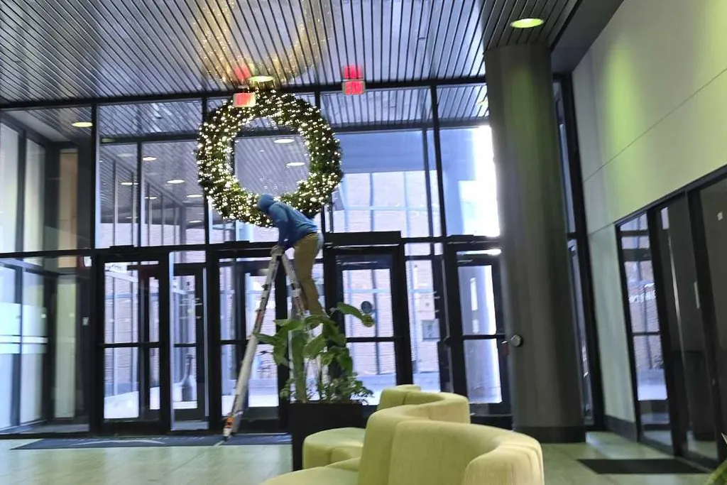 A Sharp Lawn Care installer on a stepladder hanging a large lit wreath above the glass entrance of a commercial lobby